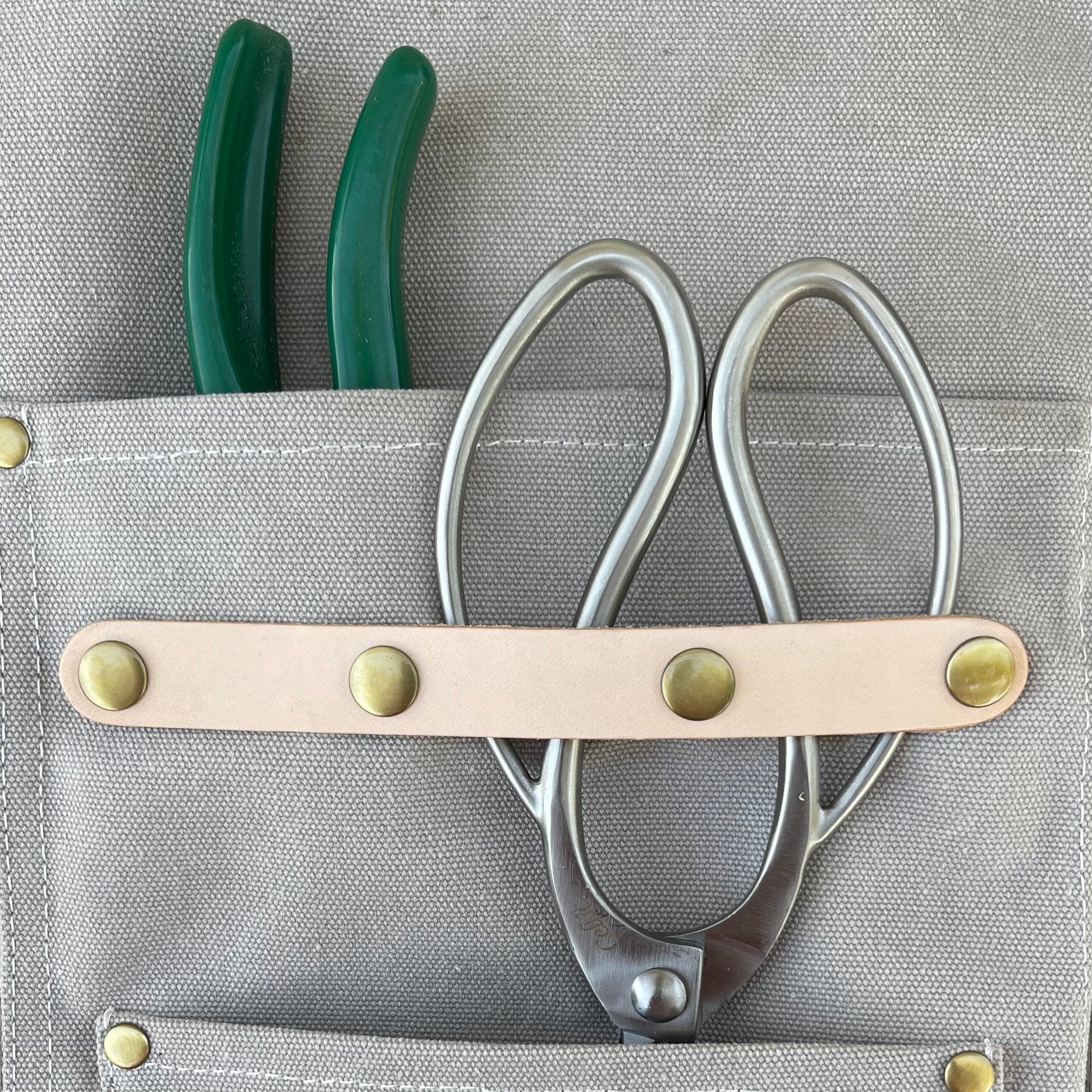 The Celtic Farm’s Gardening Apron—crafted from gray waxed canvas with pockets—holds green-handled pliers and metal scissors, secured by a beige leather strap with brass rivets; perfect for gardeners and florists.