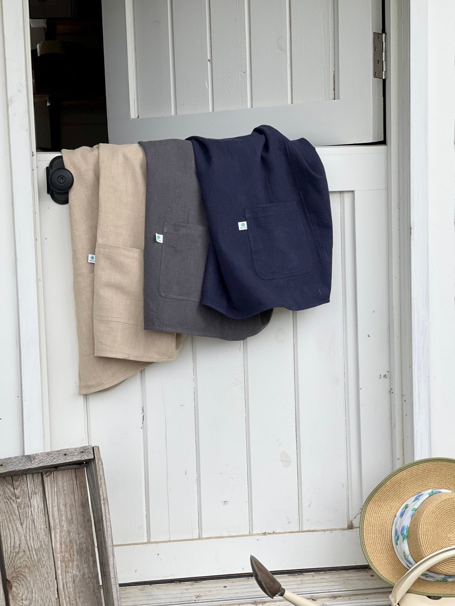 Three Linen Apron - French Style Crossback by The Celtic Farm in beige, gray, and navy hang on a white door. Nearby, a straw hat with a blue ribbon and a gardening tool evoke calm rustic charm—perfect for this multi-purpose apron.