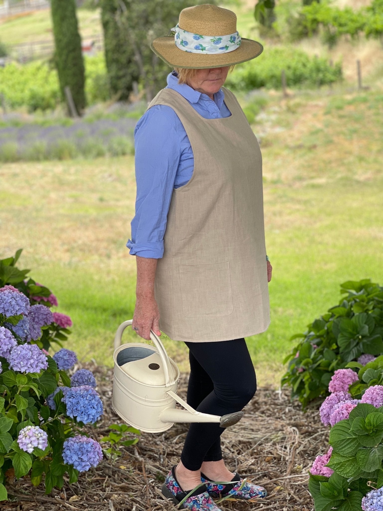 Wearing The Celtic Farm’s Linen Apron - French Style Crossback, a person in a straw hat, blue shirt, and black leggings stands outdoors among hydrangeas, holding a white watering can against a backdrop of grass and trees.