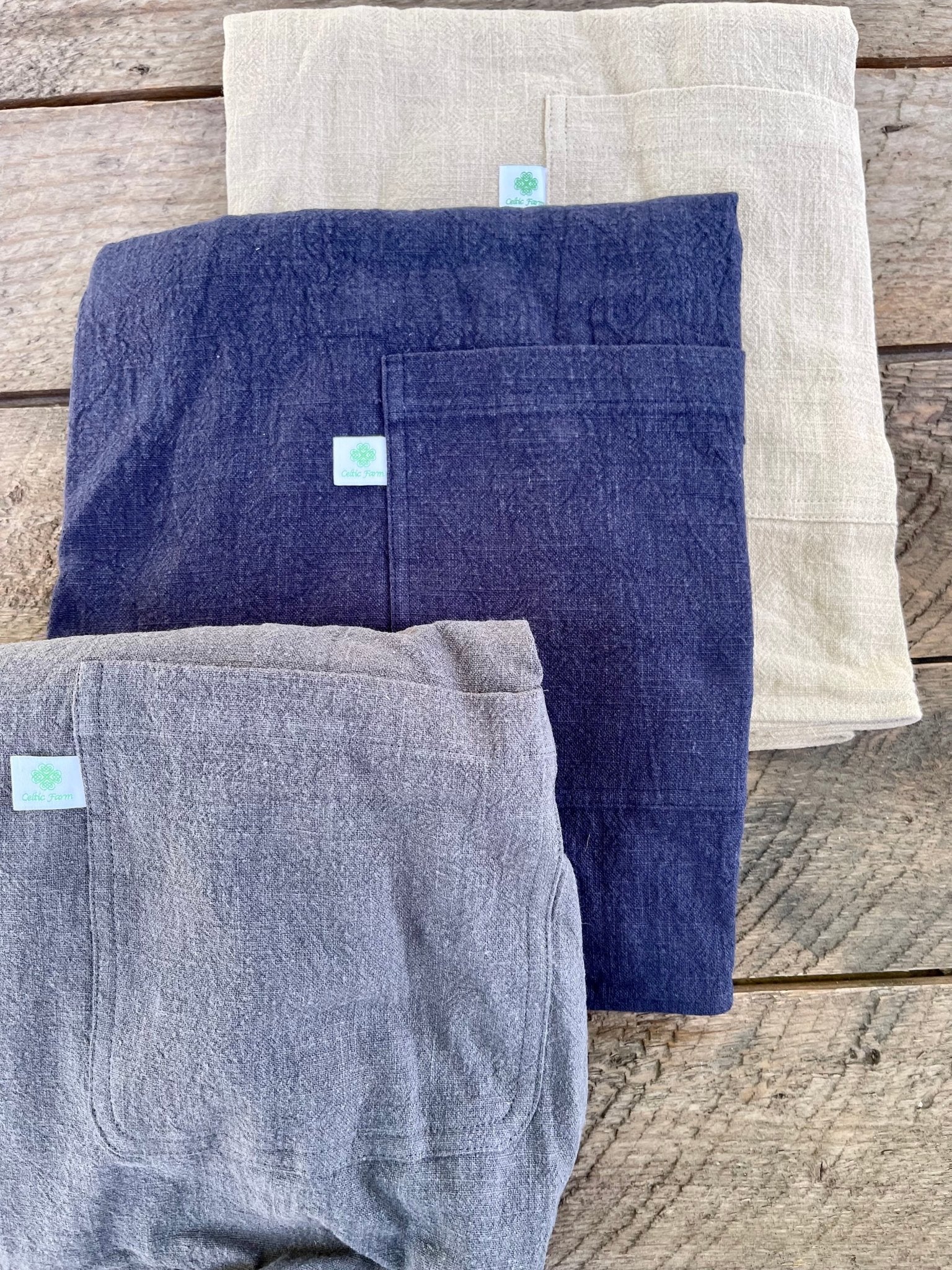 Three Linen Apron - French Style Crossback by The Celtic Farm in beige, navy blue, and gray are stacked on a rustic wooden surface. Each has a front pocket with a white tag featuring a green design.
