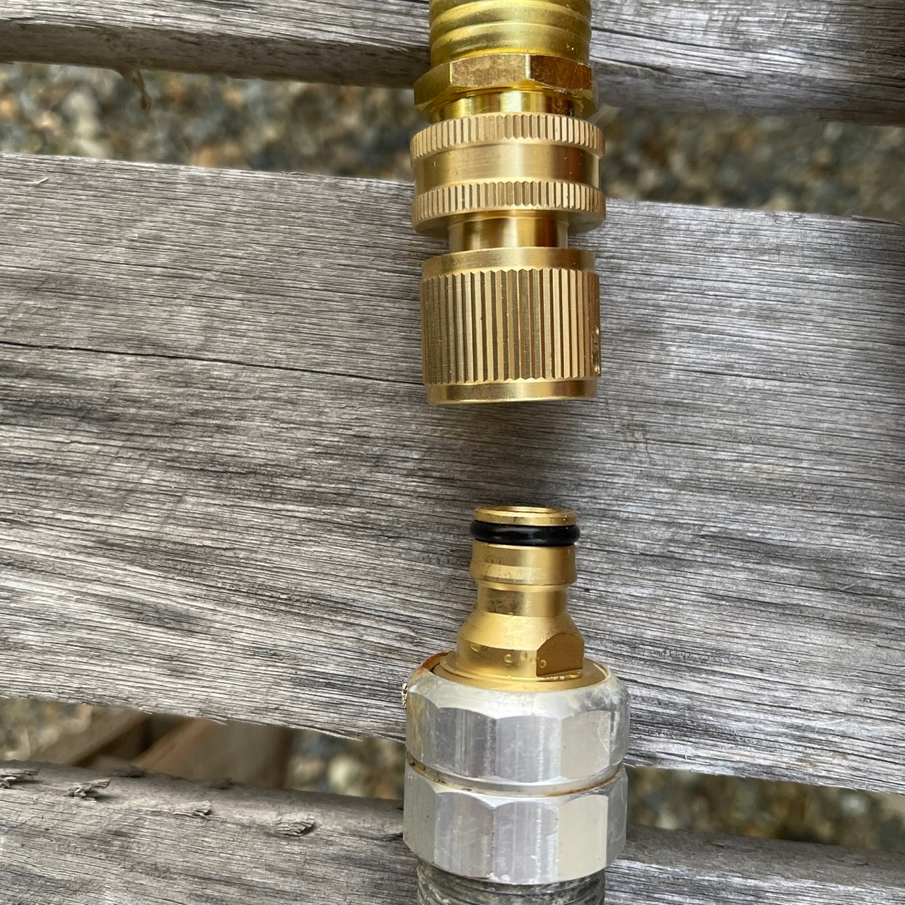 The Celtic Farm’s Brass Hose Organization Bundle—two connectors, one brass and one silver-brass—are displayed on a wooden outdoor surface with planks and gravel, ready to organize your garden hose.