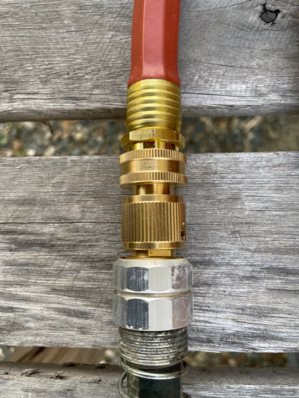 A close-up of The Celtic Farms Brass Hose Organization Bundle shows it connecting a red hose to a silver connector on a weathered wooden surface, ensuring a secure, leak-proof fit.