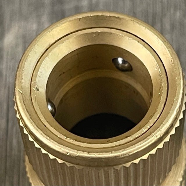 Close-up of The Celtic Farm Garden Hose Brass Quick Connect Connector featuring a threaded outer edge, circular opening, and two internal ball bearings. The background is dark and blurred.