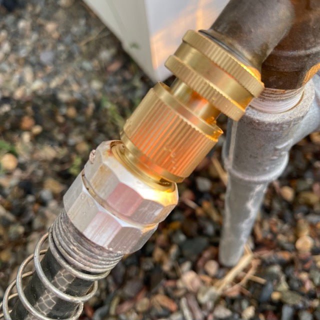 Close-up of The Celtic Farm Garden Hose Brass Quick Connect Connector secured to a pipe with a metal spring guard over the hose, ensuring a leak-proof connection above gravel and dried leaves.