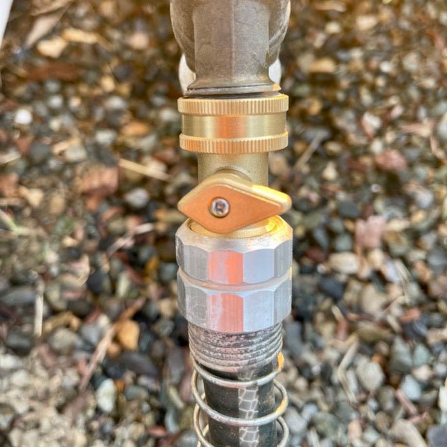Close-up of The Celtic Farm Garden Hose Brass Shutoff Valve with a yellow handle, linked to a black garden hose and silver coupler for a leak-proof connection; blurred rocks and brown leaves in the background.