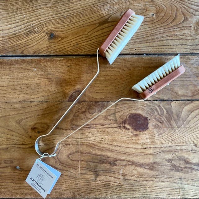 The Celtic Farms Indoor Plant Dust Brush - Unique Gift, a double-ended pearwood and goat hair brush with white bristles and curved metal handle, rests on wood. A tag is attached—ideal for gentle indoor plant care.