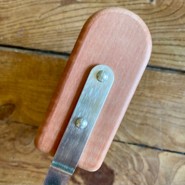 A close-up of The Celtic Farms Indoor Plant Dust Brush - Unique Gift, a rectangular pearwood tool with rounded edges and a metal strip held by two screws, resting on wood—perfect for houseplant care.