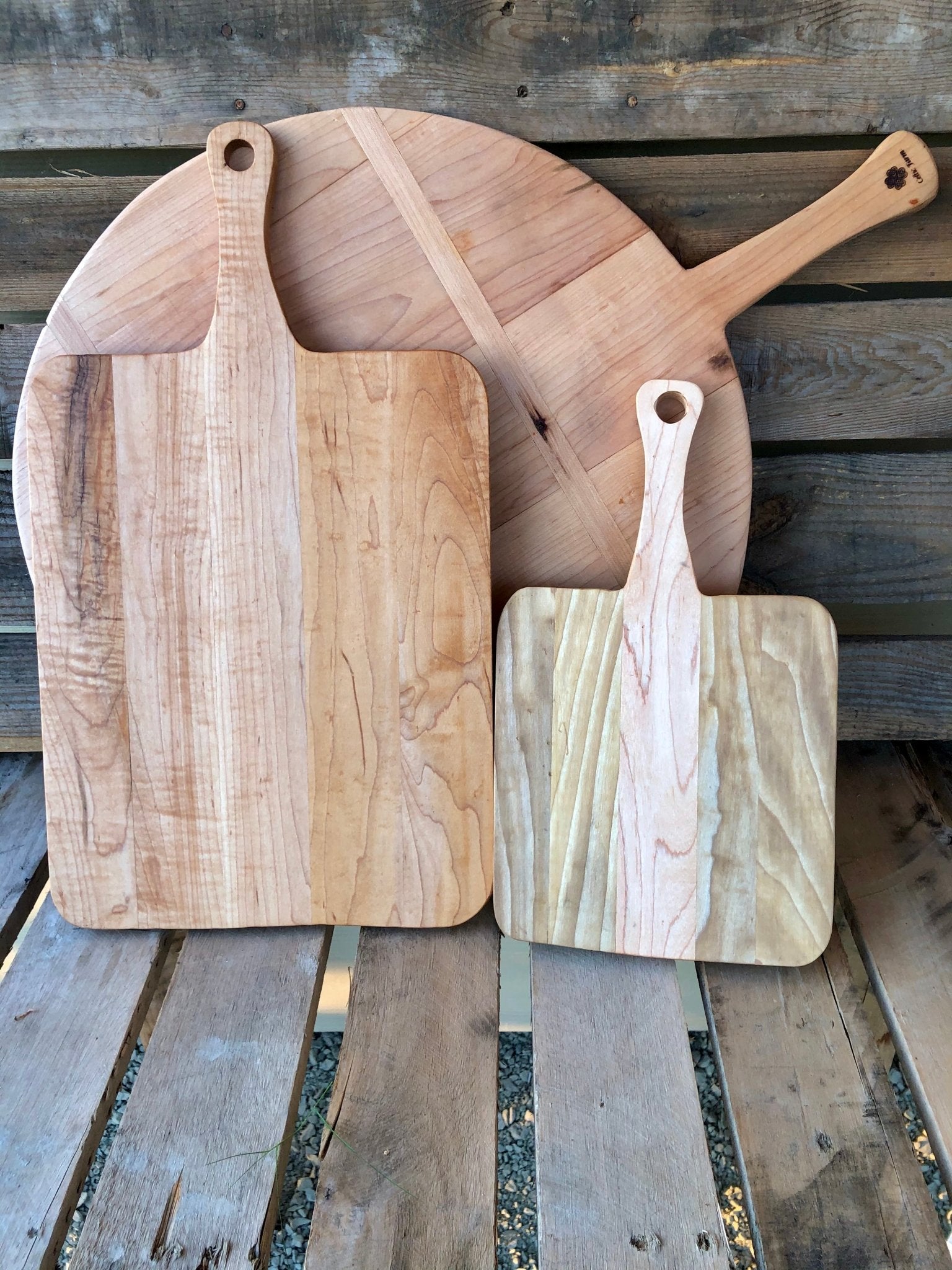Three Small Charcuterie Boards—Vintage Style Rectangle French Boards by The Celtic Farm—lean against a rustic wooden wall on a pallet, displaying natural grain and handles with hanging holes that evoke classic wood shop craftsmanship.