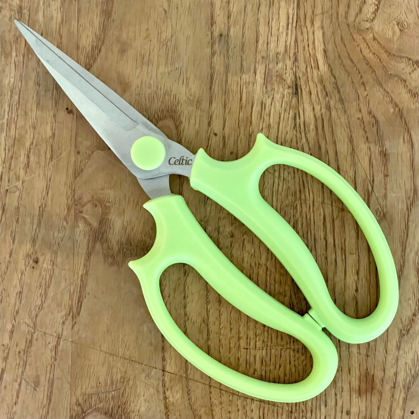 A pair of Floral & Herb Snips by The Celtic Farm, with light green handles, rests on a wooden surface. The slightly open blades are ideal for precise floral arranging and herb trimming tasks.