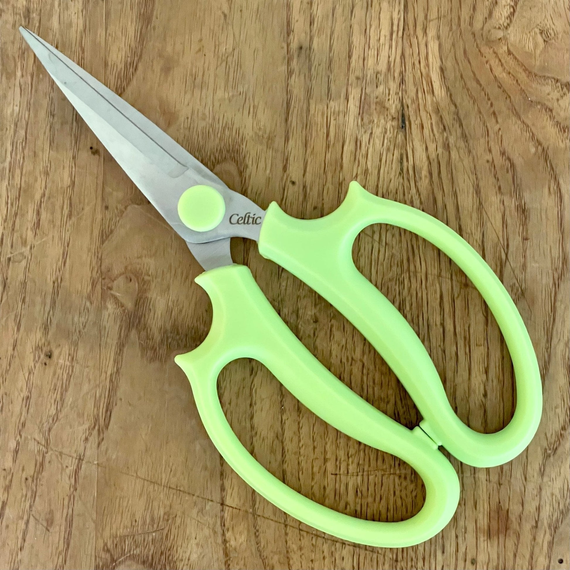 A pair of Floral & Herb Snips by The Celtic Farm, with light green handles, rests on a wooden surface. The slightly open blades are ideal for precise floral arranging and herb trimming tasks.