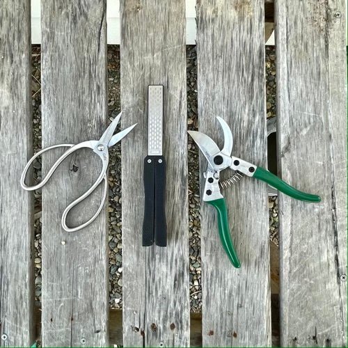 Three garden tools sit on a weathered wood surface: The Celtic Farm’s Garden Tool Sharpener with diamond + carbon steel paddle in the center, pruning shears and silver scissors nearby. Gravel is visible between the wood slats below.
