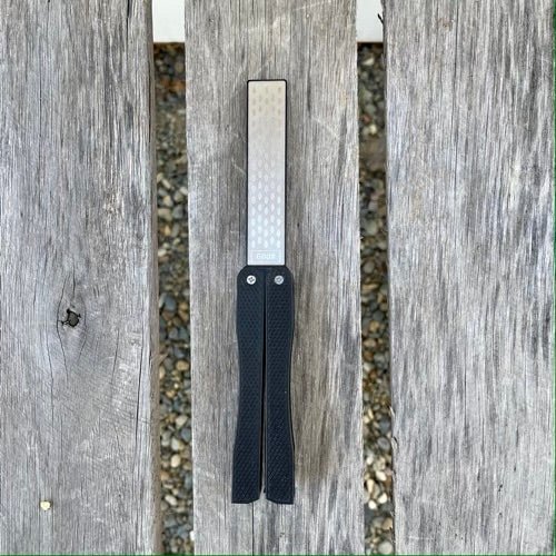 The Celtic Farm Garden Tool Sharpener, featuring a diamond and carbon steel reversible paddle, rests on weathered wood—a unique addition to your pruner, clipper, or mower blade sharpening set.