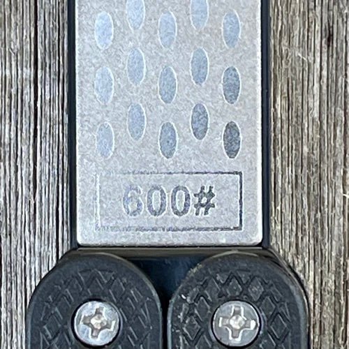 A close-up of The Celtic Farm Garden Tool Sharpener—Diamond + Carbon Steel Hone Paddle, labeled 600#, with a textured metallic finish on wood. Two black rubber grips with screws are visible at the bottom.