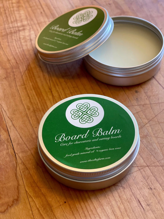 An open round tin of The Celtic Farm’s Board Balm - Oil & Wax for Cutting and Charcuterie Boards, with a green label, rests on wood. The label highlights care for boards using food-grade mineral oil and wax as a sealer and conditioner.