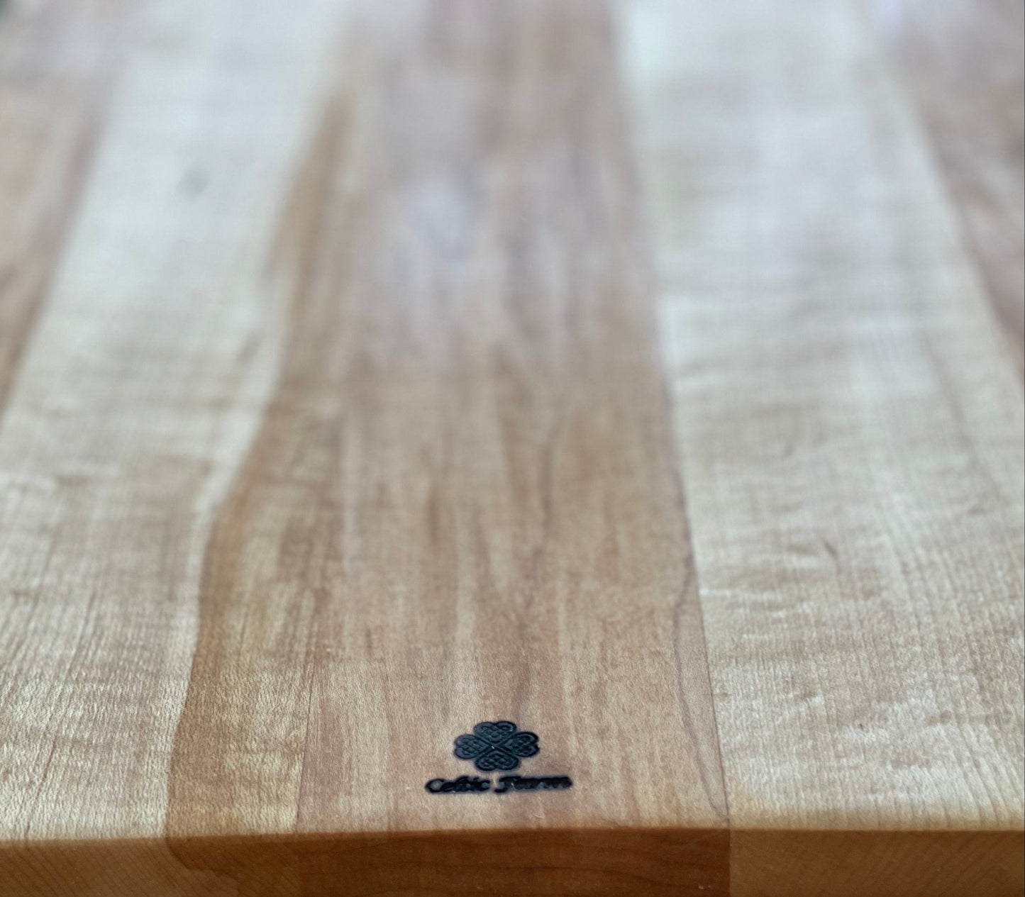 A close-up of a wooden surface conditioned with Board Balm - Oil & Wax for Cutting and Charcuterie Boards by The Celtic Farm, showing a small dark logo at the center and Celtic Fusion engraved near the wood’s edge.