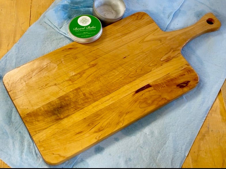 A large wooden cutting board with a handle rests on a blue cloth. Nearby are an open tin of The Celtic Farms Board Balm - Oil & Wax for Cutting and Charcuterie Boards with a green lid, and a small metal bowl.
