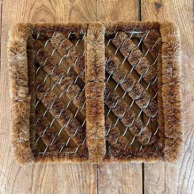 The Celtic Farms Boot Scraper Coconut Fiber Mat features coarse, natural coconut fiber bristles arranged diagonally in a wire frame with reinforced borders for durability, making it a vintage-inspired doormat perfect for scraping boots on wooden floors.