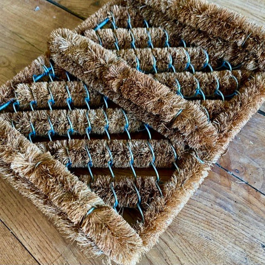 Two rectangular brown Boot Scraper Coconut Fiber Mats by The Celtic Farm, featuring stiff bristles and metal wire frames, overlap at an angle on a wooden surface. These mats are ideal for scrubbing tough dirt from footwear.