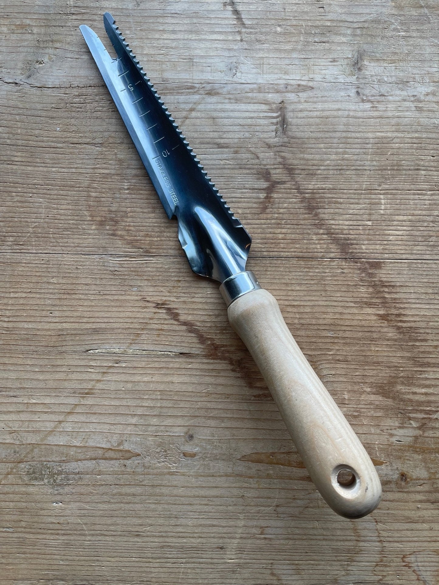 The Celtic Farms Unique Garden Tool Set & Gift features a wooden handle, serrated stainless steel blade with measurement markings, highlighted by its distinct design as it sits on a wooden surface.