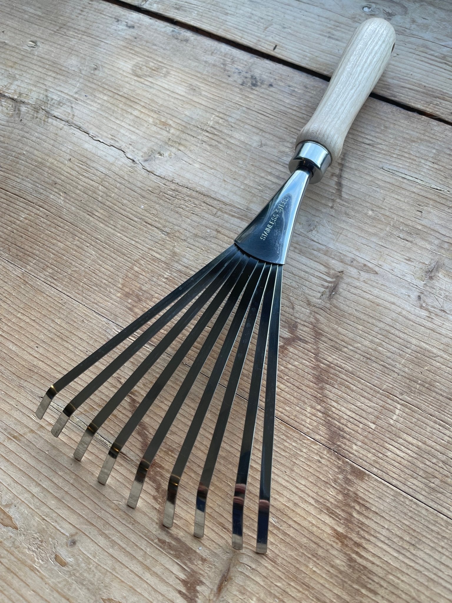 A close-up of The Celtic Farms Unique Garden Tool Set & Gift hand rake, featuring a light wooden handle and slender metal tines, resting on a wooden surface.