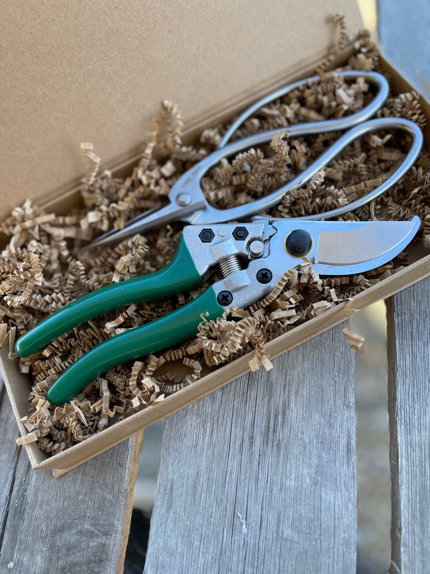 The Celtic Farm Garden Gift Box - Pruners and Snips features green-handled tools nestled in brown crinkle paper, elegantly presented on a rustic wooden surface.