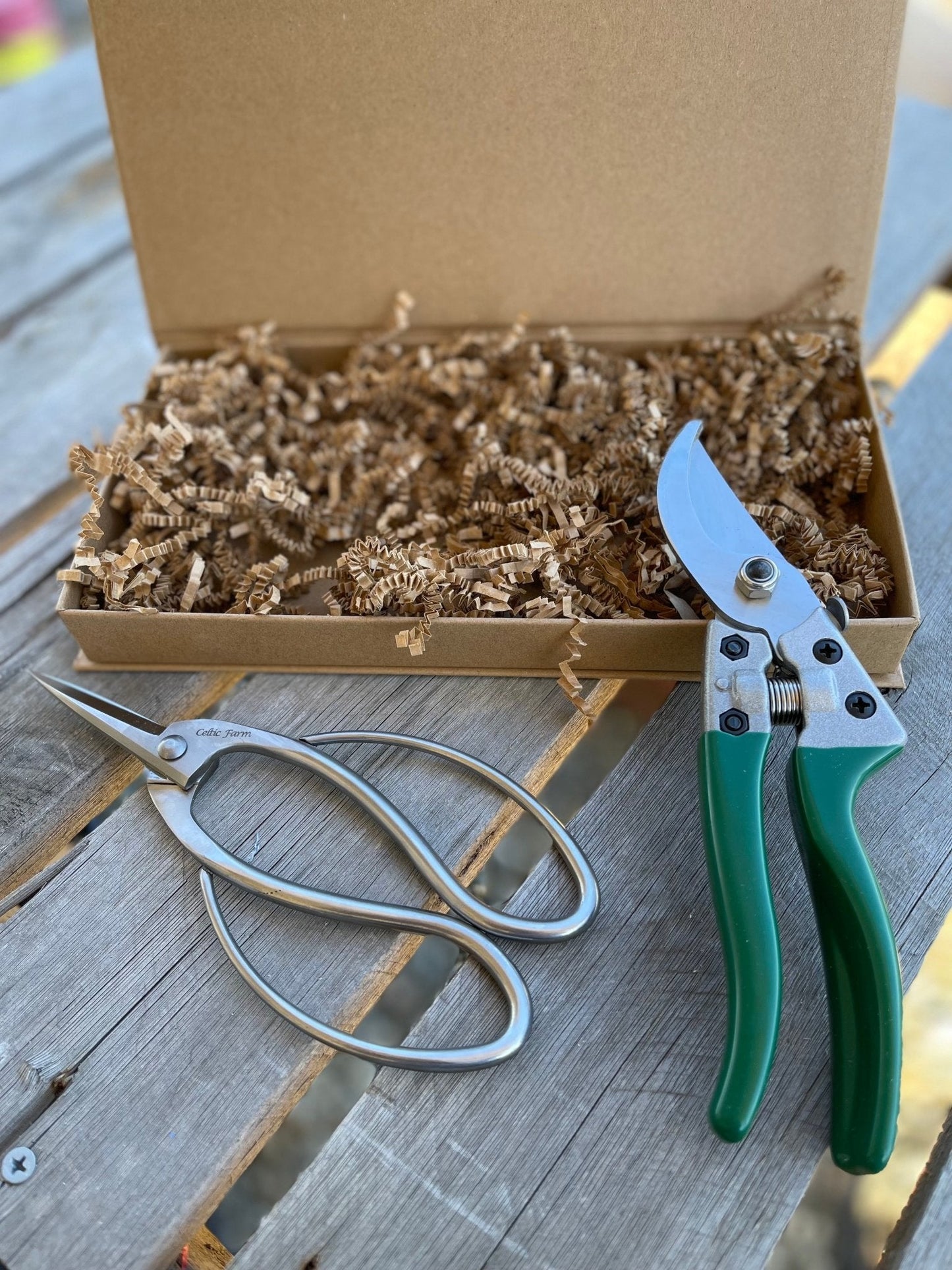 Two pairs of pruners from The Celtic Farm’s Garden Gift Box—one green-handled, one silver—rest on wood beside the open box, which is filled with brown crinkle paper.