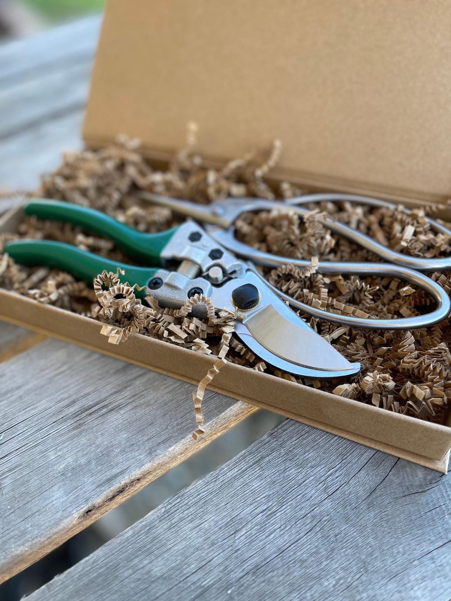 The Celtic Farm’s Garden Gift Box - Pruners and Snips features green-handled pruners nestled in brown crinkle paper inside an open box, displayed on a rustic wooden surface.