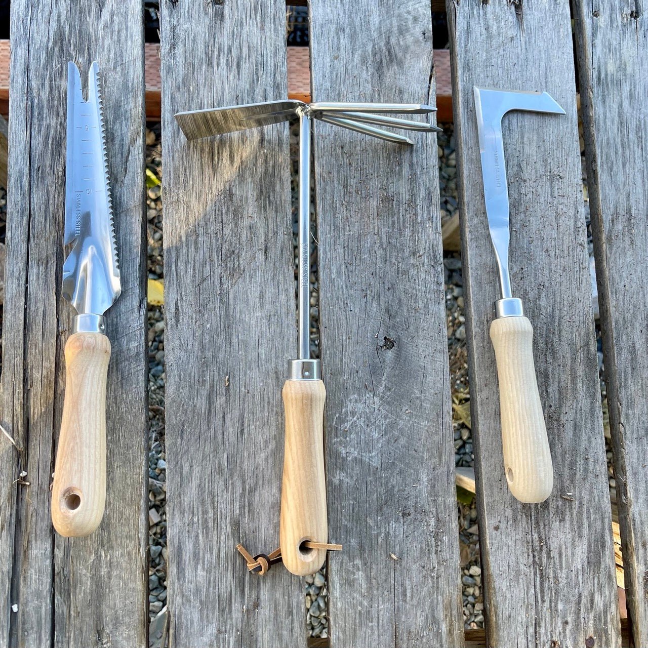 The Celtic Farm’s Unique Garden Tool Set & Gift Two features a Ground Breaker soil knife, a three-prong cultivator, and a hooked blade weeder, each with wooden handles, displayed on a rustic wood surface.