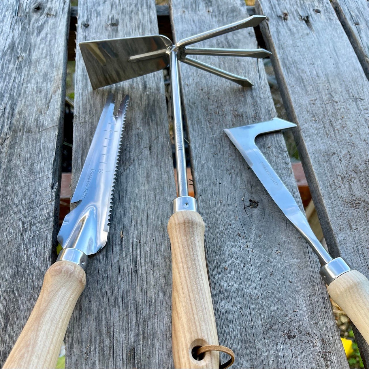 The Celtic Farms Unique Garden Tool Set & Gift Two features three metal tools with wooden handles—a serrated trowel, a three-pronged cultivator, and a Ground Breaker weeder—displayed on a weathered wooden surface.