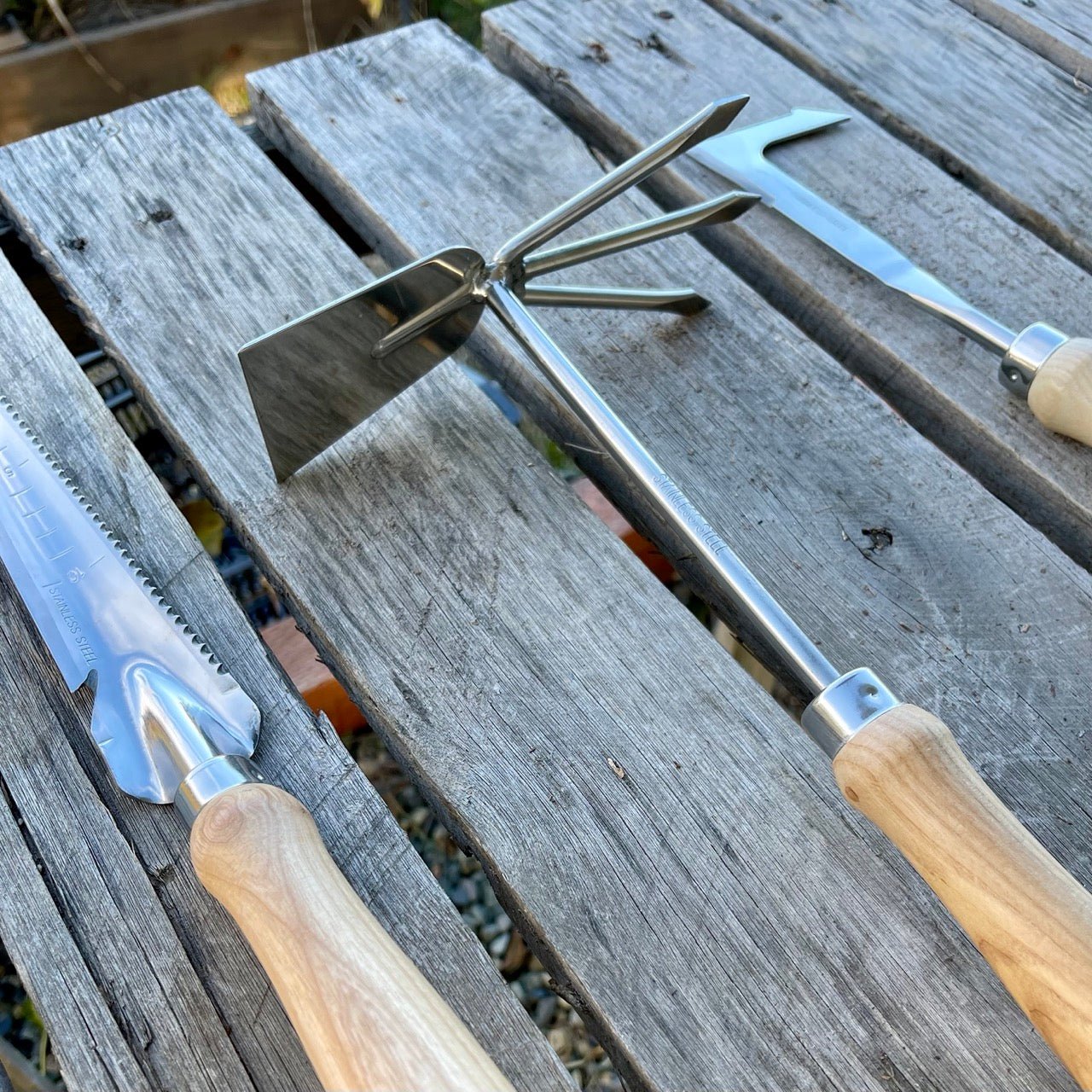 The Celtic Farms Unique Garden Tool Set & Gift Two features three stainless steel garden tools with wooden handles—a Ground Breaker trowel, weeder, and flat-edged tool—displayed on a weathered wooden table outdoors.