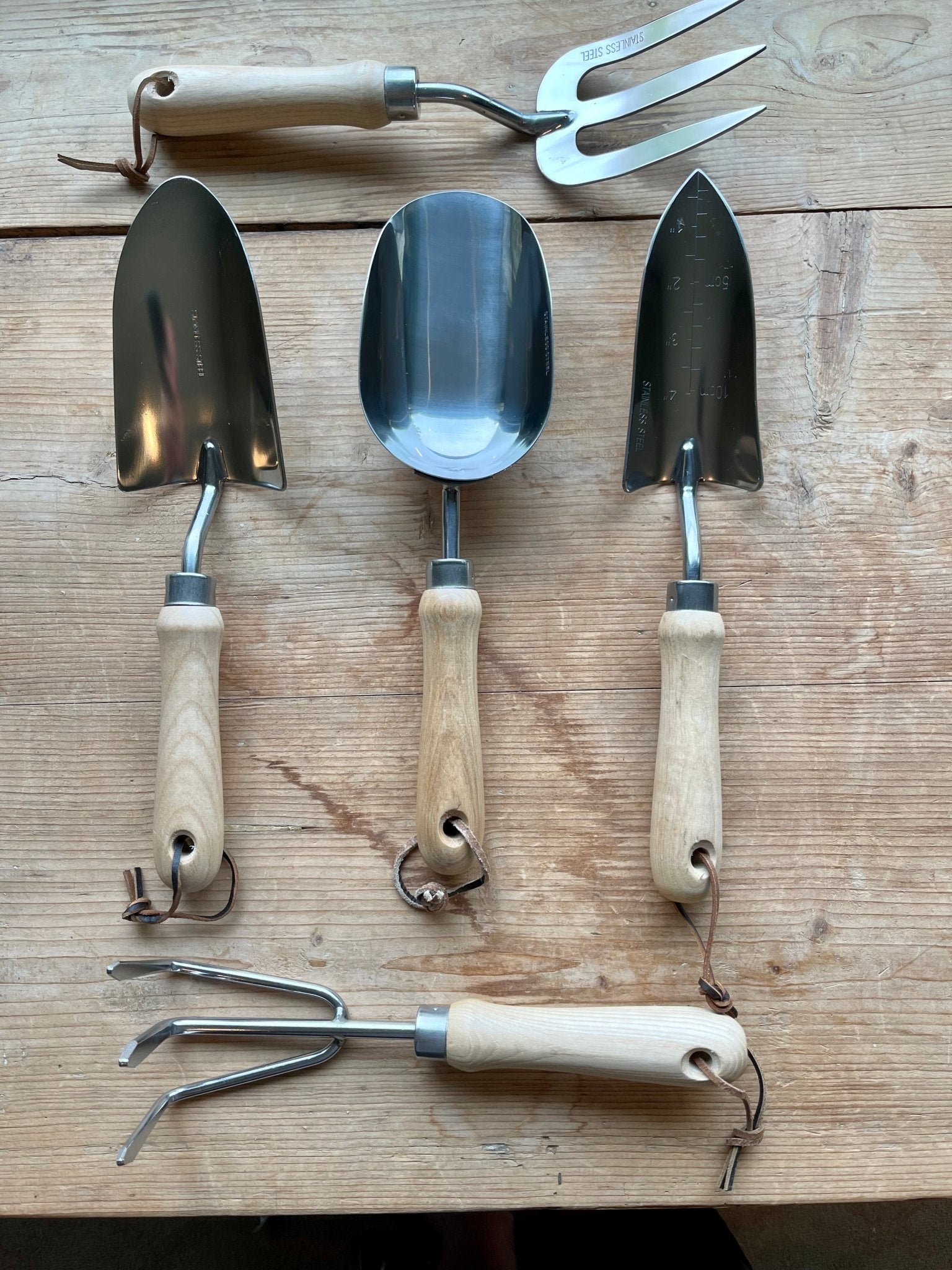 The Celtic Farms Garden Master Tool Set features five wooden-handled tools—two trowels, a scoop, a weeder, and a fork—displayed on wood, showcasing their stainless finish and classic craftsmanship.