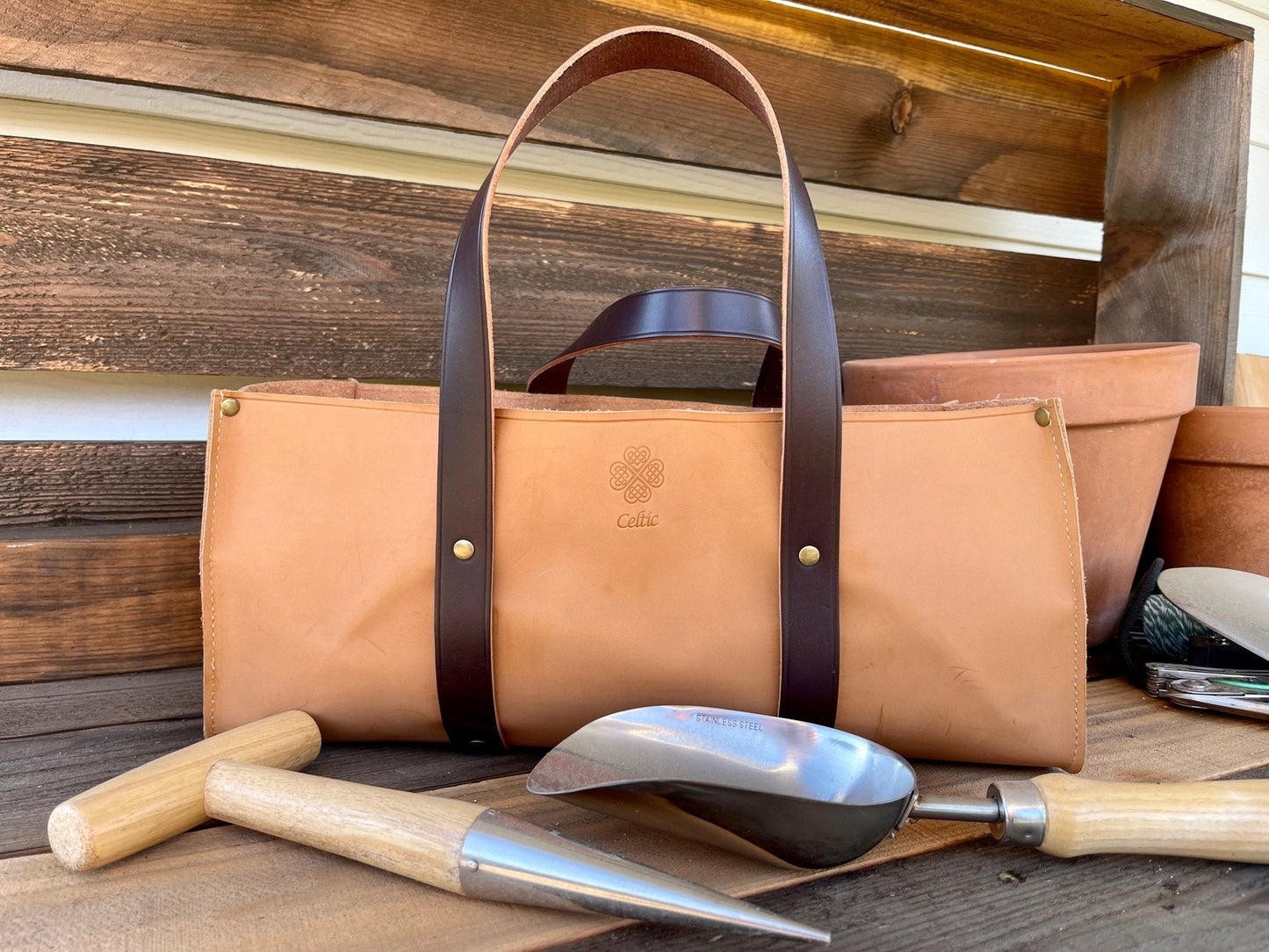 A tan leather Garden Master Tool Set bag by The Celtic Farm, featuring dark brown handles and a Celtic knot logo, sits on wood with stainless and hardwood tools and terra cotta pots. The word Celtic is embossed on the bag.