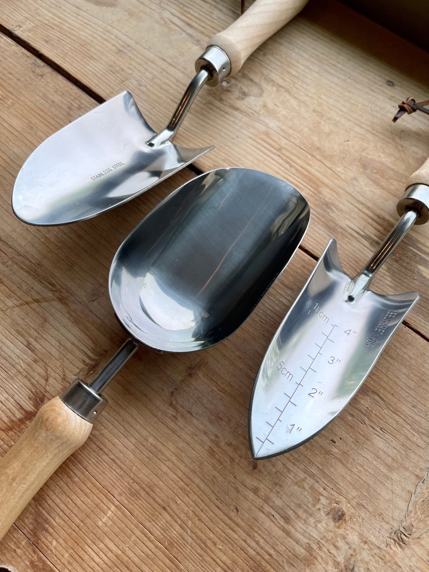 The Celtic Farm Potters Tool Set includes three metal trowels with wooden handles—one with measurement markings and one wide scoop—resting on wood, ideal for use with a Burlap Garden Bag as essential potting tools.
