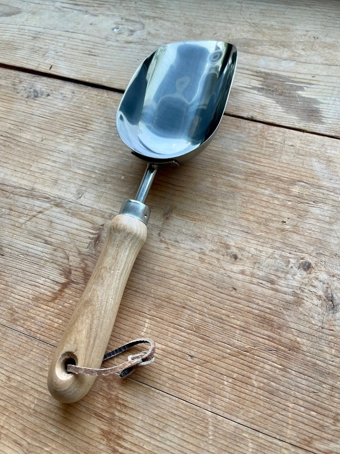 A metal garden scoop with a wooden handle and leather strap from The Celtic Farms Potters Tool Set - 3 Tools for Potting rests on rustic wood, making it a perfect Garden Tool Gift or addition to any potting toolkit.
