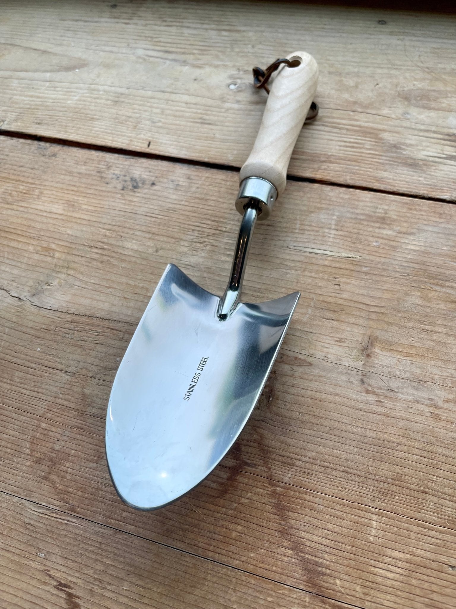 A shiny metal trowel with a light wooden handle from The Celtic Farms Potters Tool Set - 3 Tools for Potting rests on wood, making it a perfect Garden Tool Gift for any enthusiast.