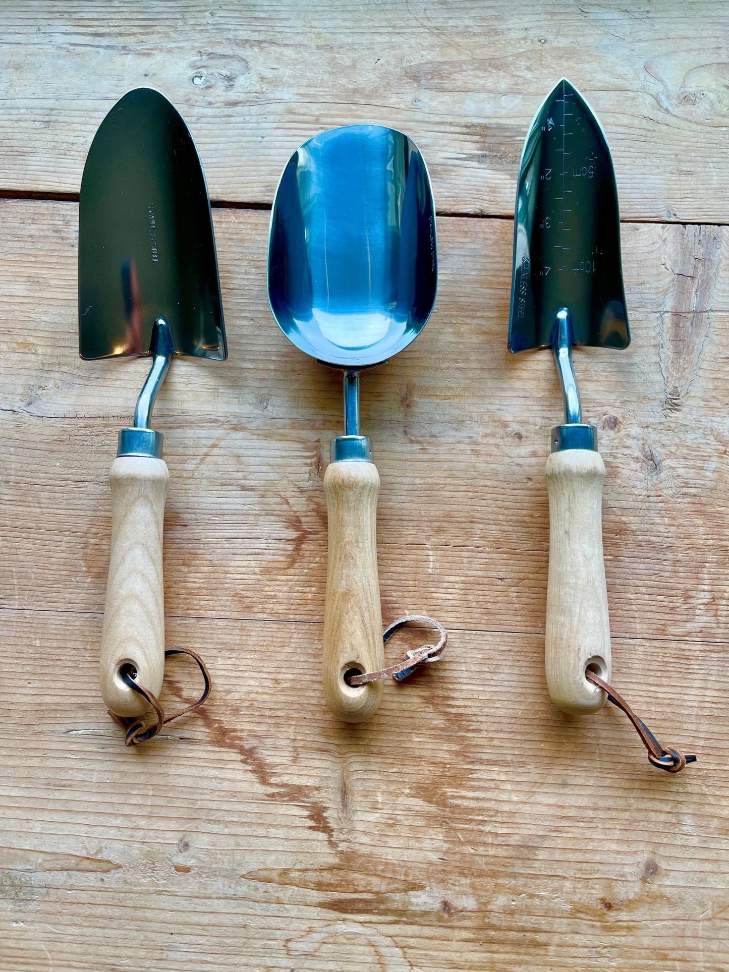 The Celtic Farm’s Potters Tool Set includes three potting tools with wooden handles, metal heads, and leather loops—two trowels and a scoop—side by side on wood. Ideal as a Garden Tool Gift.