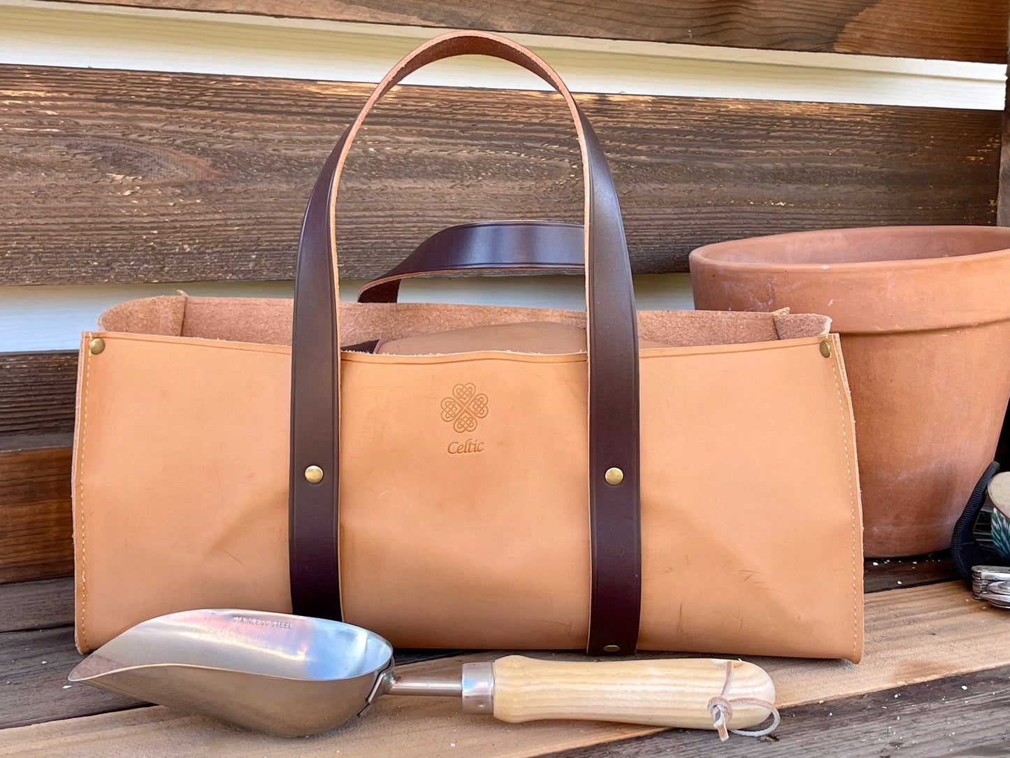 The Celtic Farms Potters Tool Set – 3 Tools for Potting is displayed before stacked terracotta pots on a wooden surface, with a metal scoop featuring a wooden handle resting in front.