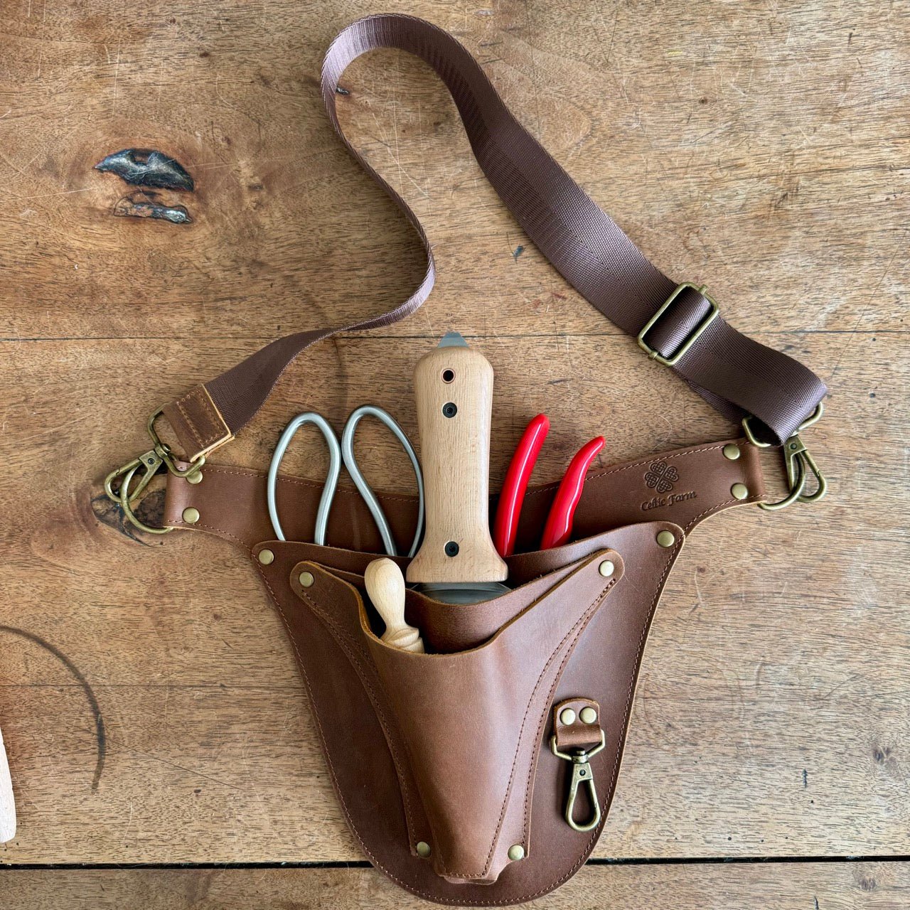The Celtic Farm Ultimate Leather Garden Tool Belt & Holder, extra rivet reinforced and master gardener designed, organizes knives, pruning shears, red-handled clippers, and wooden-handled tools with a handy shoulder strap.