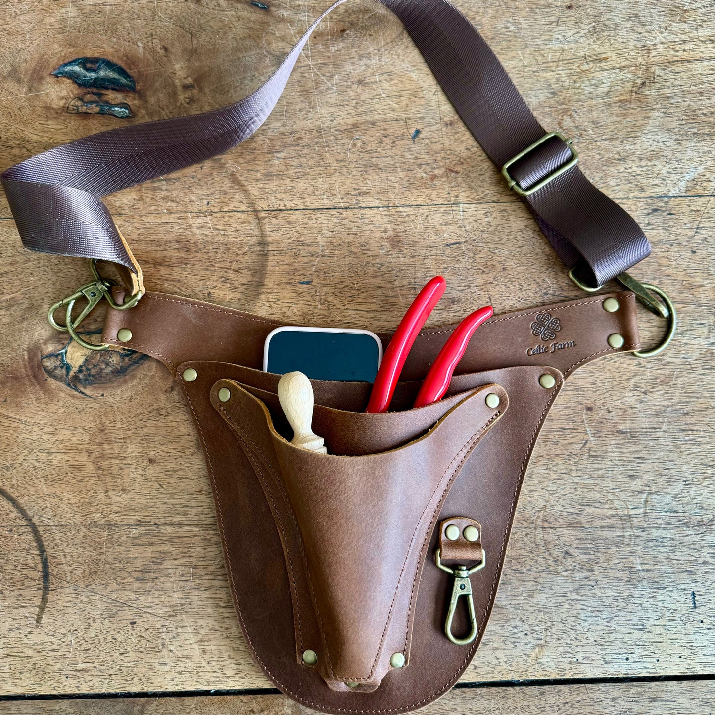 The Celtic Farms Ultimate Leather Garden Tool Belt & Holder—extra rivet reinforced, master gardener designed—securely holds your phone, wooden dibber, and two red-handled pliers for essential tool organization on any surface.