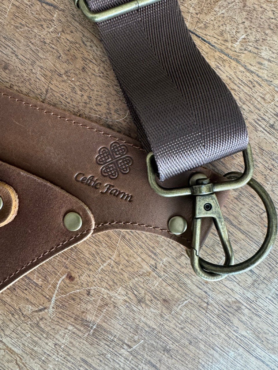 A close-up of The Celtic Farm’s Ultimate Leather Garden Tool Belt & Holder, featuring extra-rivet reinforcement, metal hardware, and a Celtic knot embossing, shown on a worn wood surface. Designed by master gardeners for your favorite tools.