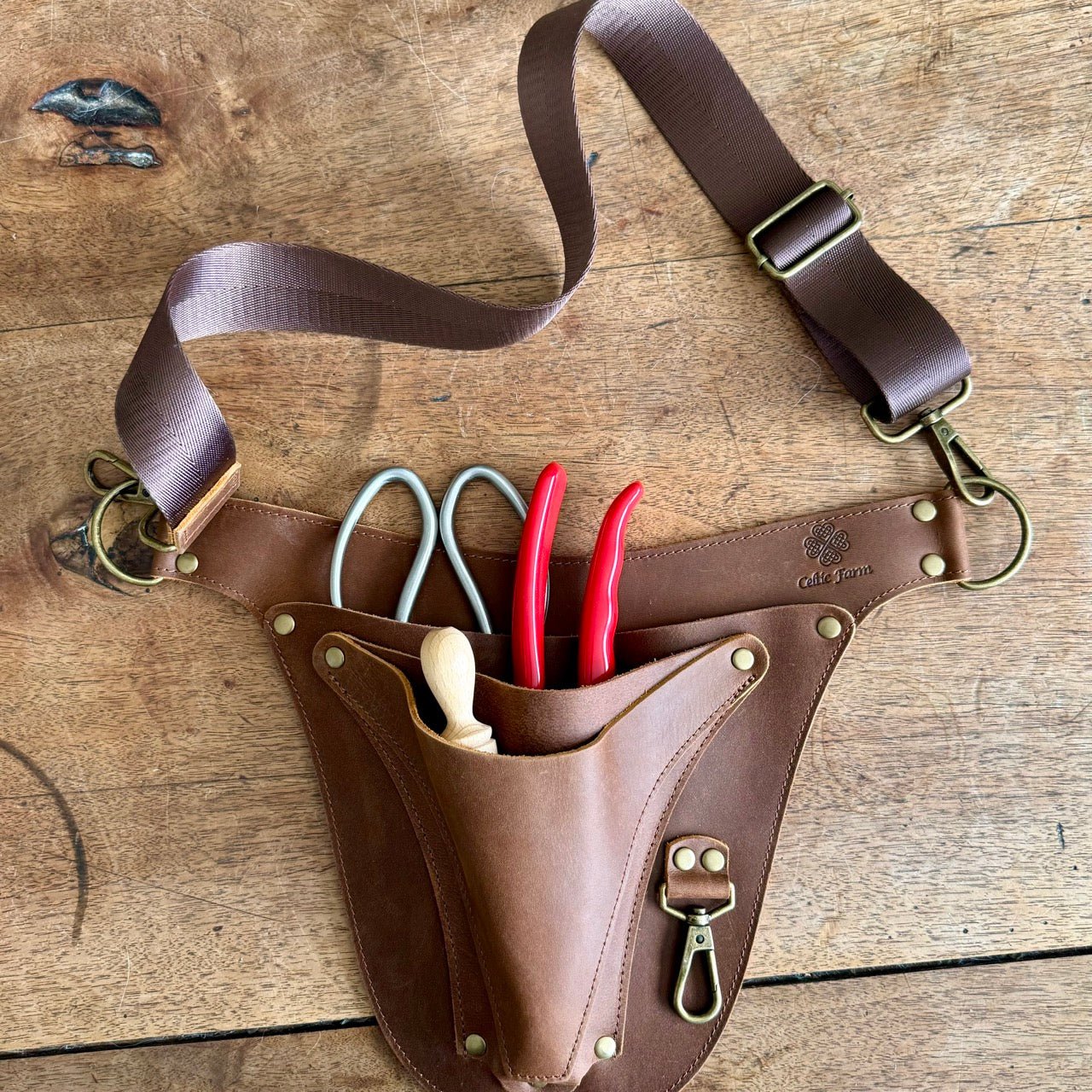 The Celtic Farm’s Ultimate Leather Garden Tool Belt & Holder, master gardener designed and extra rivet reinforced, sits on a wooden surface holding two pairs of scissors, red-handled pliers, and a wooden gardening tool.