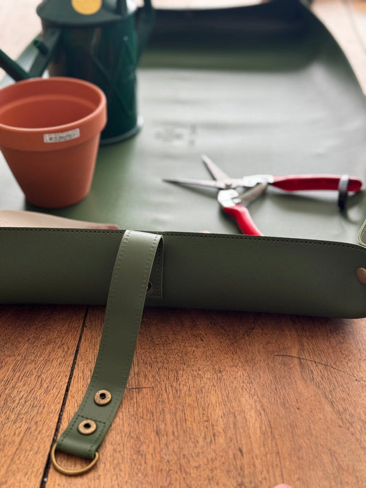 A The Celtic Farm Potting Mat - Waterproof For Tidy Gardening, pruning shears, a terracotta pot, and a green watering can sit on a wooden table with a gardening tool bag—perfect for compact storage and easy cleanup.