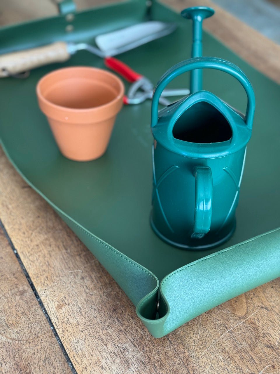 On a wooden table, The Celtic Farm’s Potting Mat - Waterproof For Tidy Gardening holds a terracotta flowerpot, green plastic watering can, red-handled garden shears, and a trowel with a wooden handle.