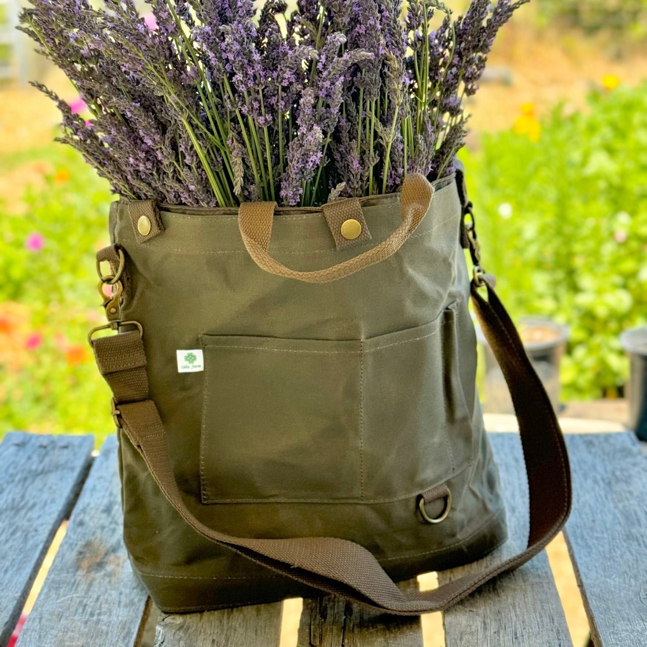 The Celtic Farm Gardeners Multipurpose Shoulder Bag in green waxed canvas with brown straps sits on a wooden table, filled with purple lavender stems. A garden with blurred greenery and flowers is in the background.