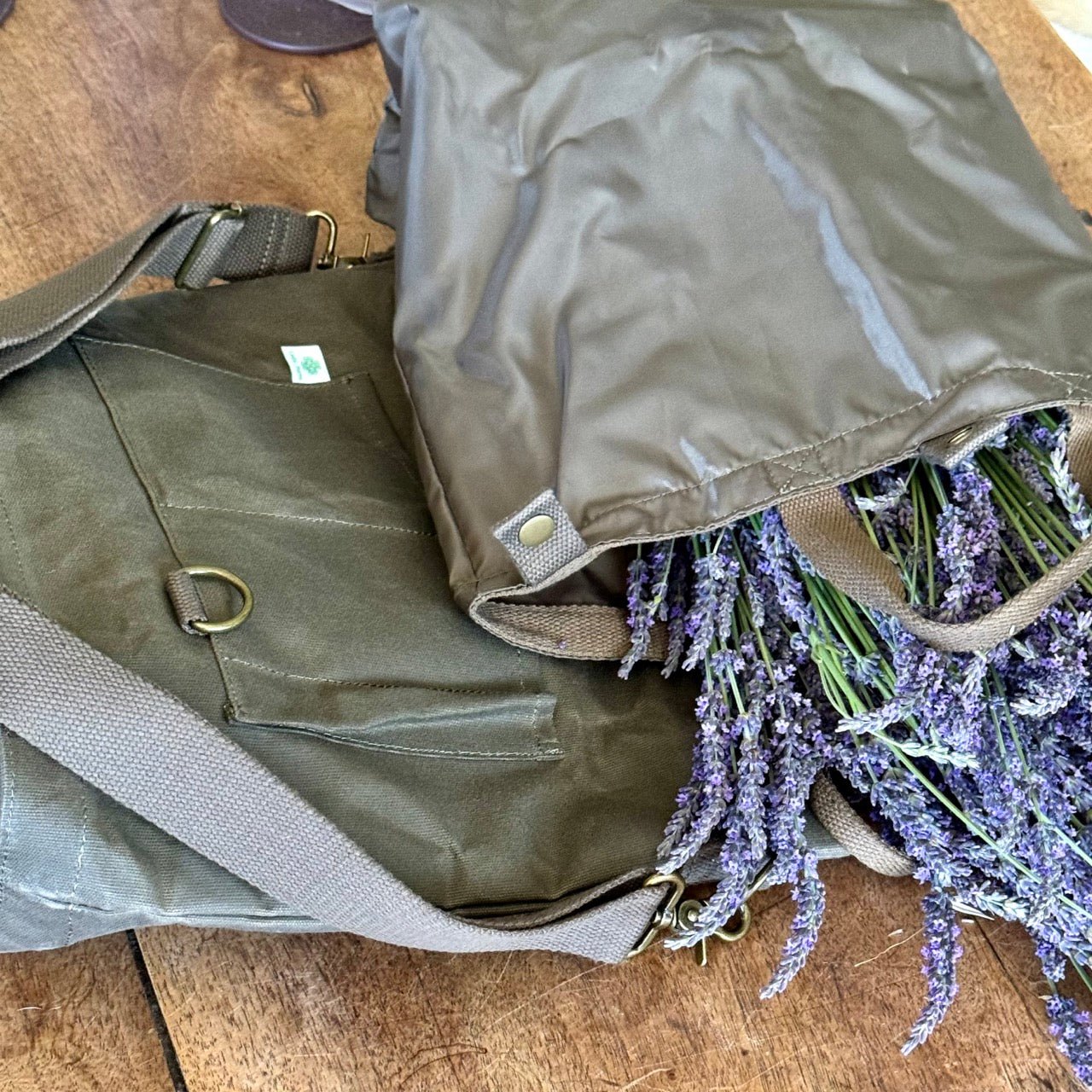 Two Gardeners Multipurpose Shoulder Bags by The Celtic Farm rest on wood. One open bag reveals fresh purple lavender bunches. Both have beige shoulder straps and metal clasps for organized tool, market, or harvest storage.