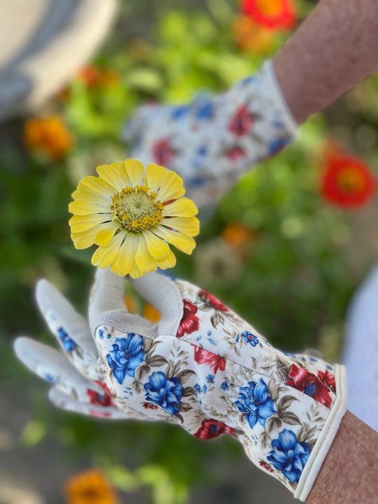 A person wearing The Celtic Farms Womens Floral Gardening and Project Gloves The Caroline holds a yellow flower against a blurred background of green plants and orange blooms.