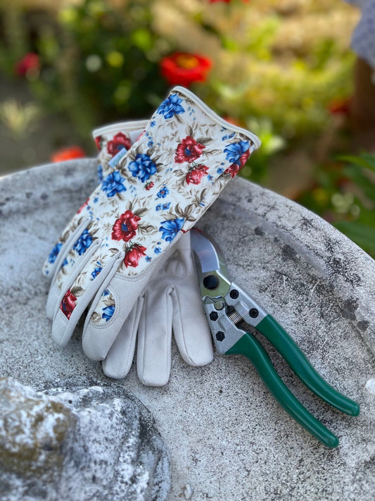 A pair of The Celtic Farms The Caroline Womens Floral Gardening and Project Gloves rests beside green-handled pruning shears on a round stone surface, with blurred greenery and red flowers in the background.