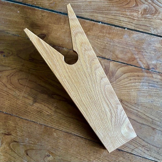 The Celtic Farm’s English Boot Jack - White Oak features a wedge shape, two prongs, and a central round hole—ideal for removing garden boots. It rests on a wooden floor in the photo.
