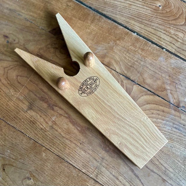 The Celtic Farm’s English Boot Jack - White Oak rests on a wooden floor, showcasing two pegs, a V-shaped notch, and a vintage-style label—perfect for easily removing garden boots.