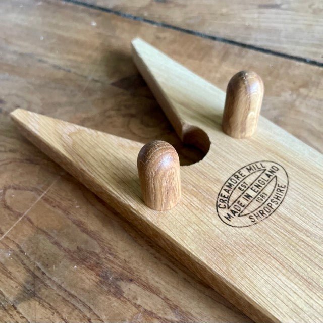 The Celtic Farm’s English Boot Jack - White Oak features two upright pegs and a “Creamore Mill Made in England Shropshire” stamp, making it perfect for removing garden boots. It rests elegantly on a wooden surface.